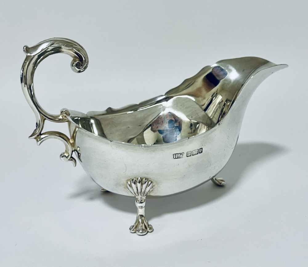 A polished antique silver sauce boat featuring a wavy scalloped rim, a double scroll handle, and three shell-capped cabriole feet. View IV