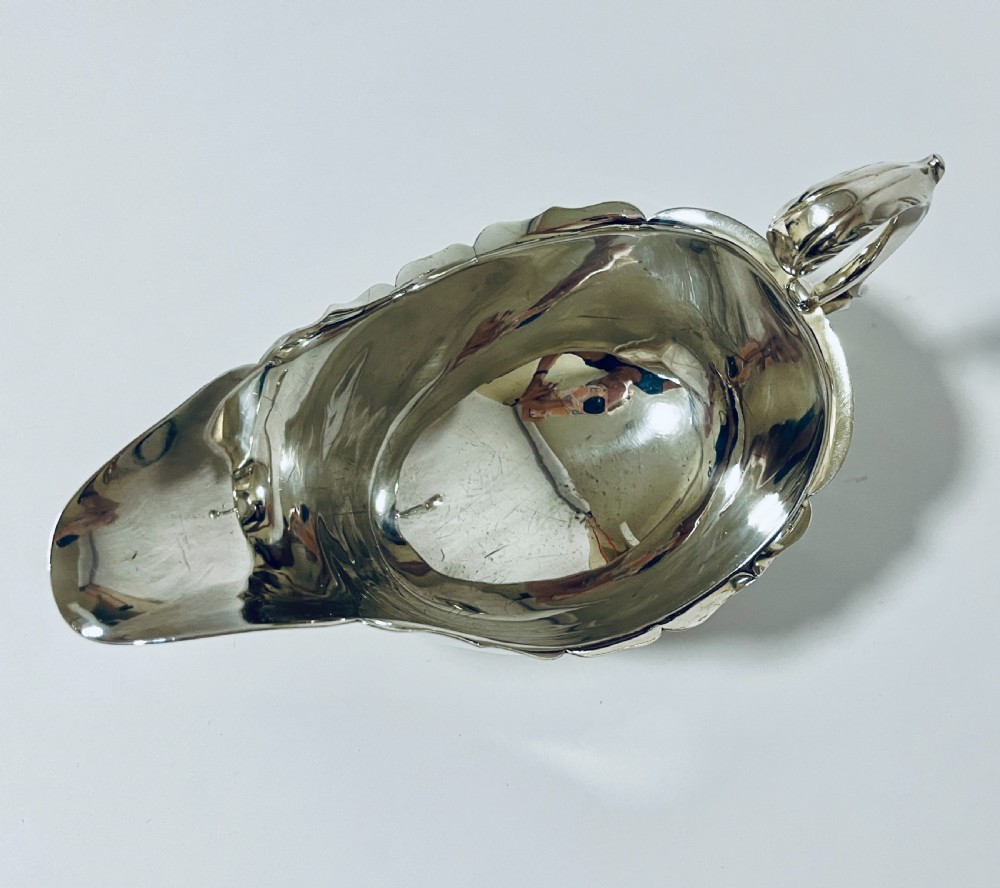 A polished antique silver sauce boat featuring a wavy scalloped rim, a double scroll handle, and three shell-capped cabriole feet. View VII
