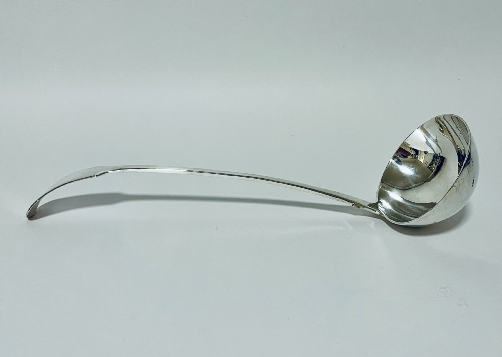 A polished Georgian silver serving ladle featuring a deep circular bowl and a long curved handle resting on a plain light surface.