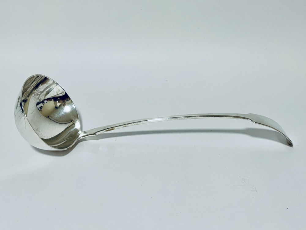 A polished Georgian silver serving ladle featuring a deep circular bowl and a long curved handle resting on a plain light surface. View II