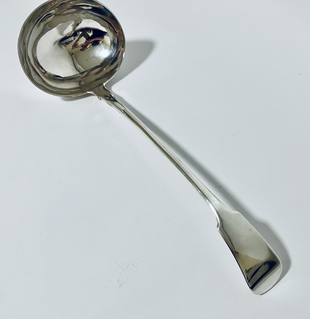 A polished Georgian silver serving ladle featuring a deep circular bowl and a long curved handle resting on a plain light surface. View III