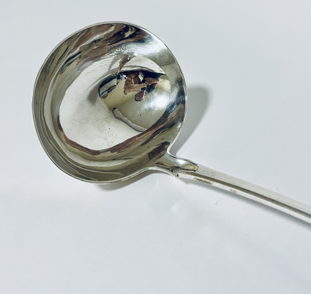 A polished Georgian silver serving ladle featuring a deep circular bowl and a long curved handle resting on a plain light surface. View IV