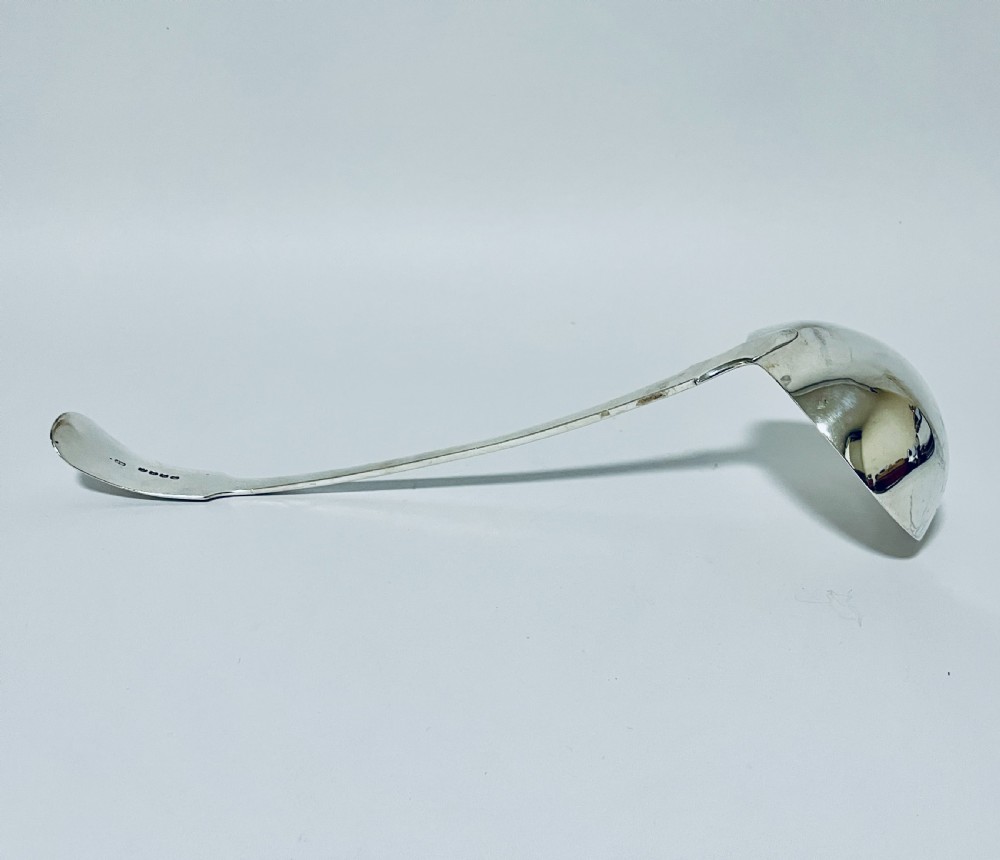 A polished Georgian silver serving ladle featuring a deep circular bowl and a long curved handle resting on a plain light surface. View V