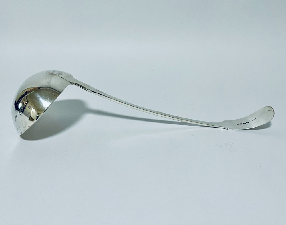 A polished Georgian silver serving ladle featuring a deep circular bowl and a long curved handle resting on a plain light surface. View VI
