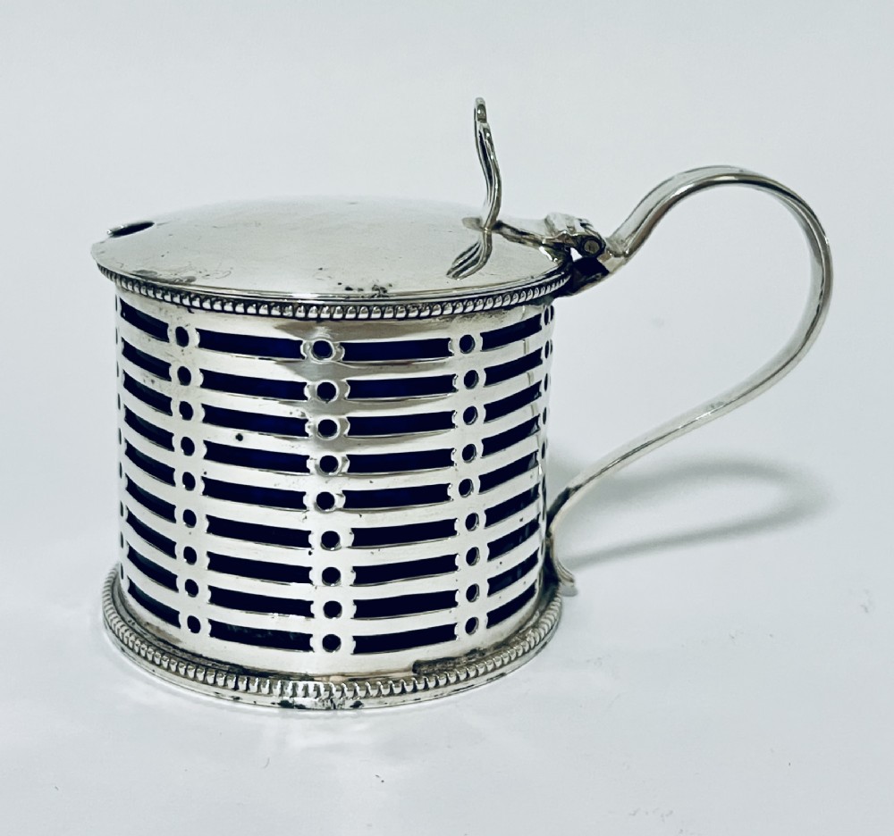 A cylindrical pierced silver mustard pot featuring a hinged lid, side handle, beaded borders, and a cobalt blue glass liner.