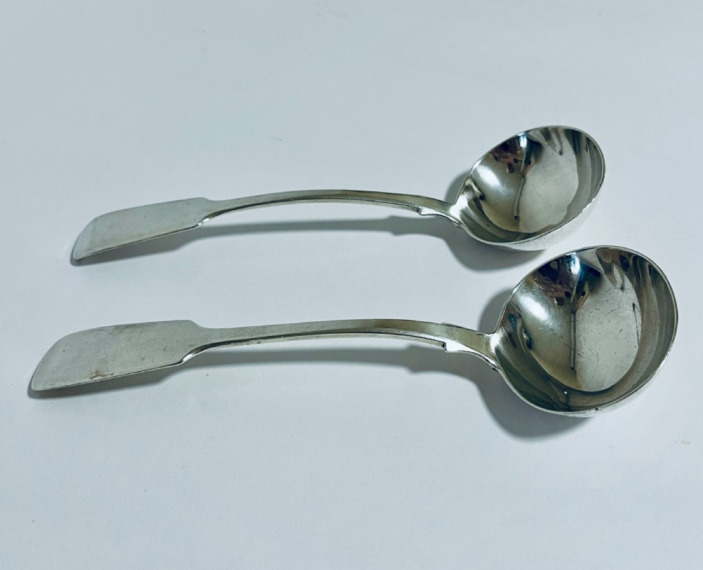 A pair of Georgian sterling silver sauce ladles with fiddle pattern handles and deep circular bowls arranged on a plain surface.