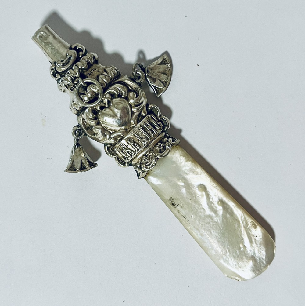 An antique silver baby rattle and teether featuring a flat mother of pearl handle, suspended silver bells, an integrated whistle top, and embossed lettering spelling DARLING.