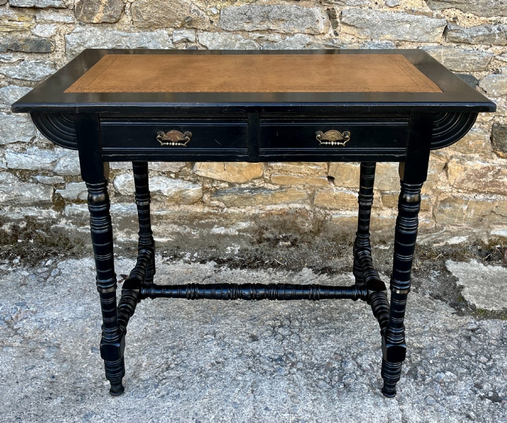 An Aesthetic Movement ebonised writing desk with a tan leather inset top, two frieze drawers with brass handles, and turned spindle legs.