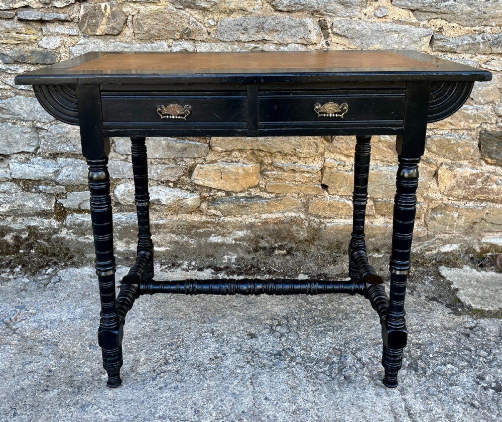 An Aesthetic Movement ebonised writing desk with a tan leather inset top, two frieze drawers with brass handles, and turned spindle legs. View II