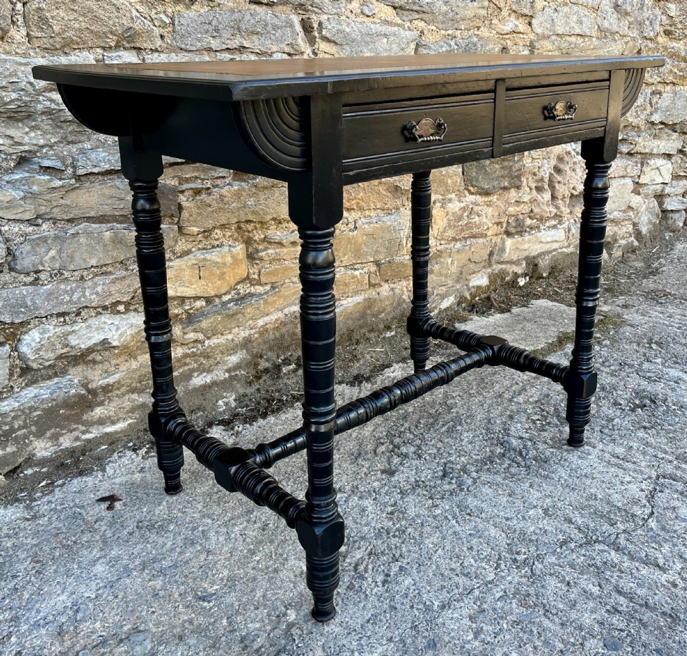 An Aesthetic Movement ebonised writing desk with a tan leather inset top, two frieze drawers with brass handles, and turned spindle legs. View V