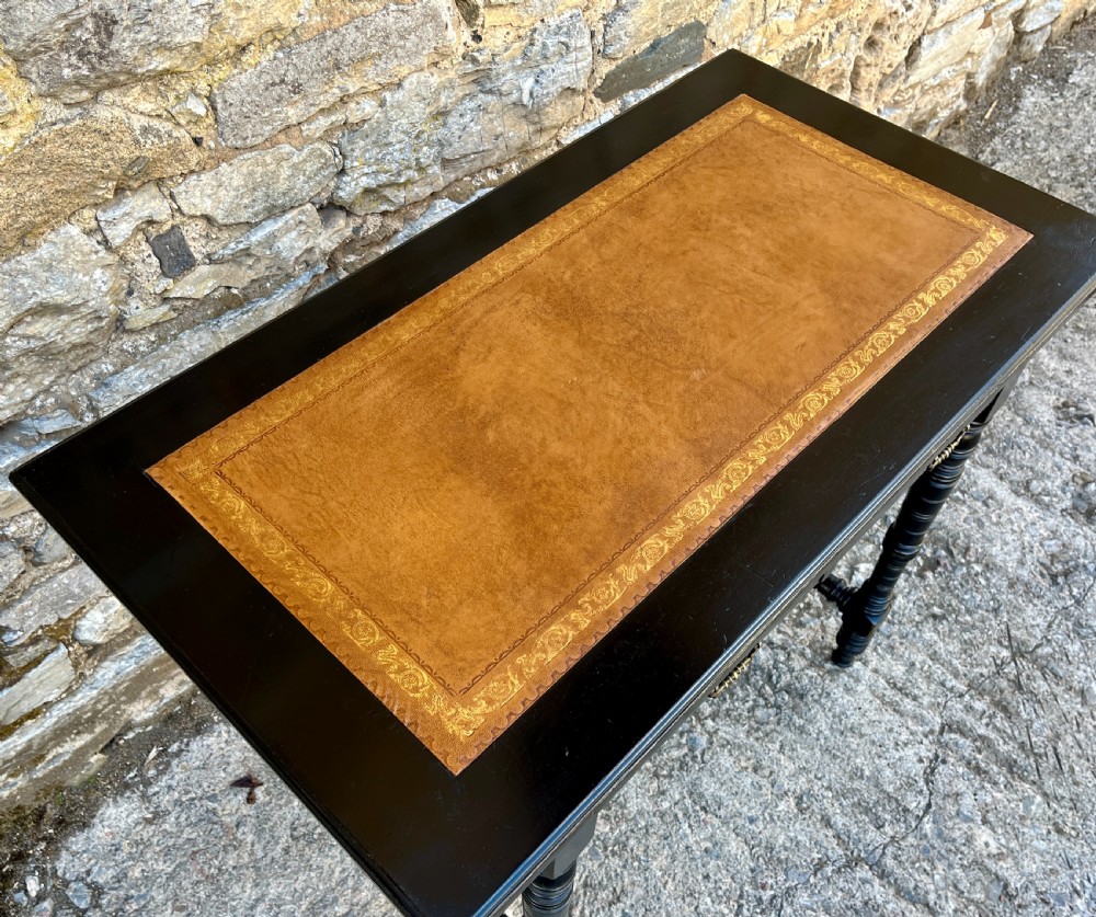 An Aesthetic Movement ebonised writing desk with a tan leather inset top, two frieze drawers with brass handles, and turned spindle legs. View IX