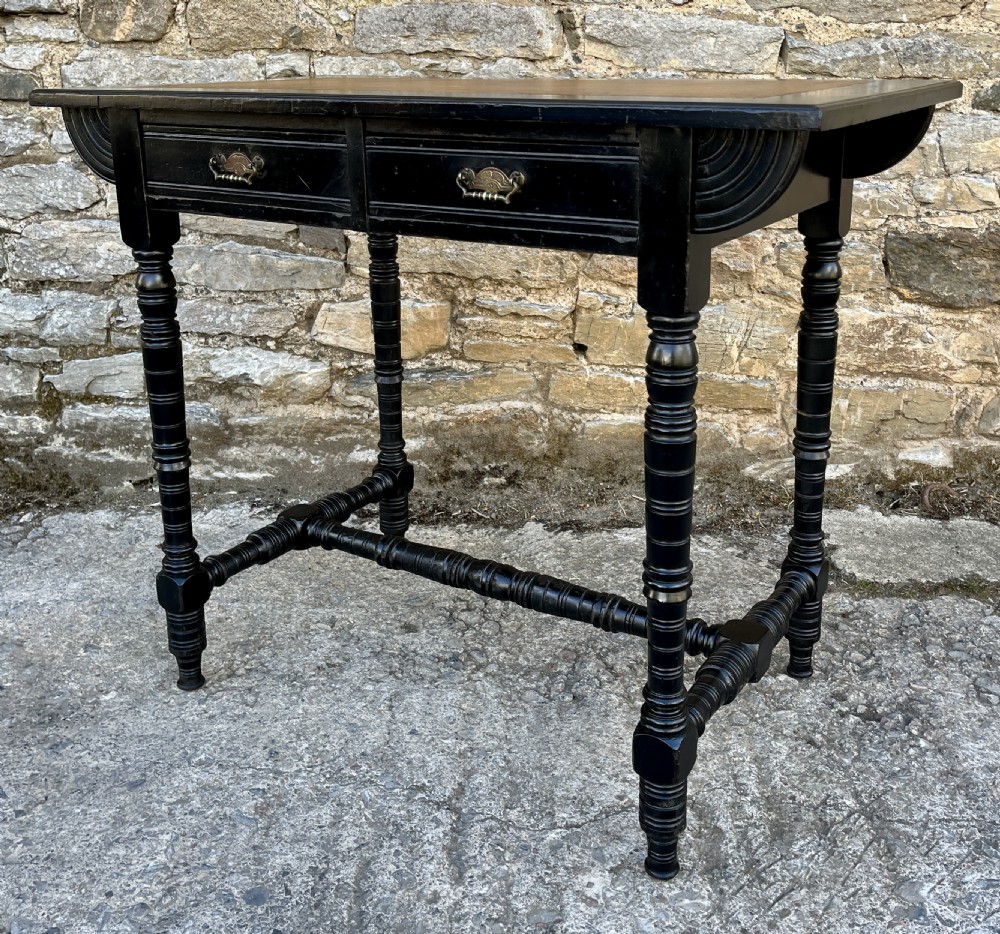 An Aesthetic Movement ebonised writing desk with a tan leather inset top, two frieze drawers with brass handles, and turned spindle legs. View XV