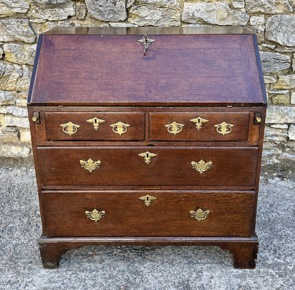An antique dark oak Georgian bureau featuring a slant front, ornate brass escutcheons, lopers, and bracket feet.