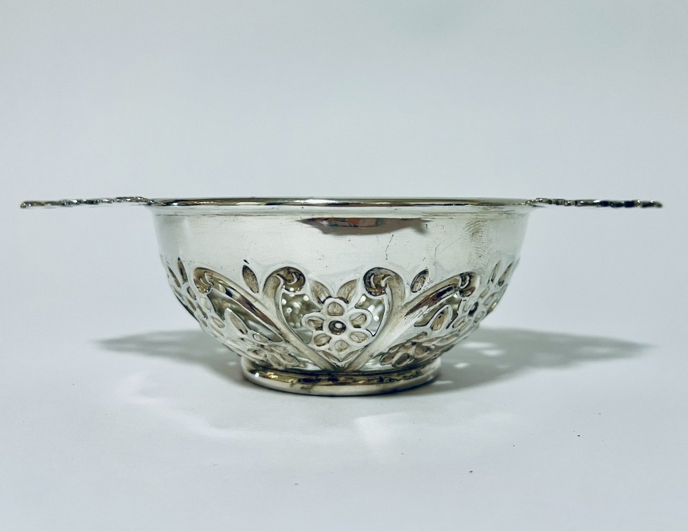 An Edwardian sterling silver quaich featuring intricate pierced floral sides, two ornate cast scroll handles, and hallmarks visible on the bowl. View VI