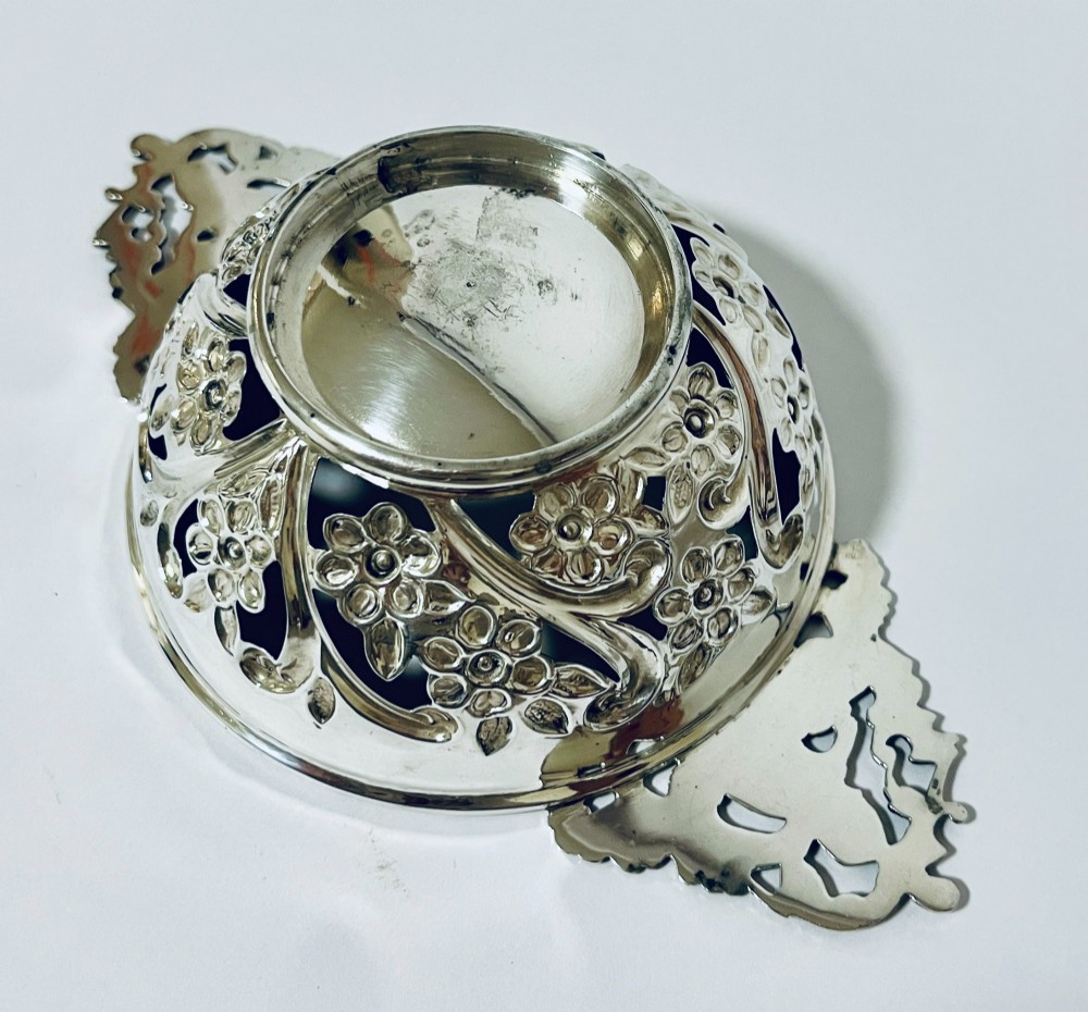 An Edwardian sterling silver quaich featuring intricate pierced floral sides, two ornate cast scroll handles, and hallmarks visible on the bowl. View IX