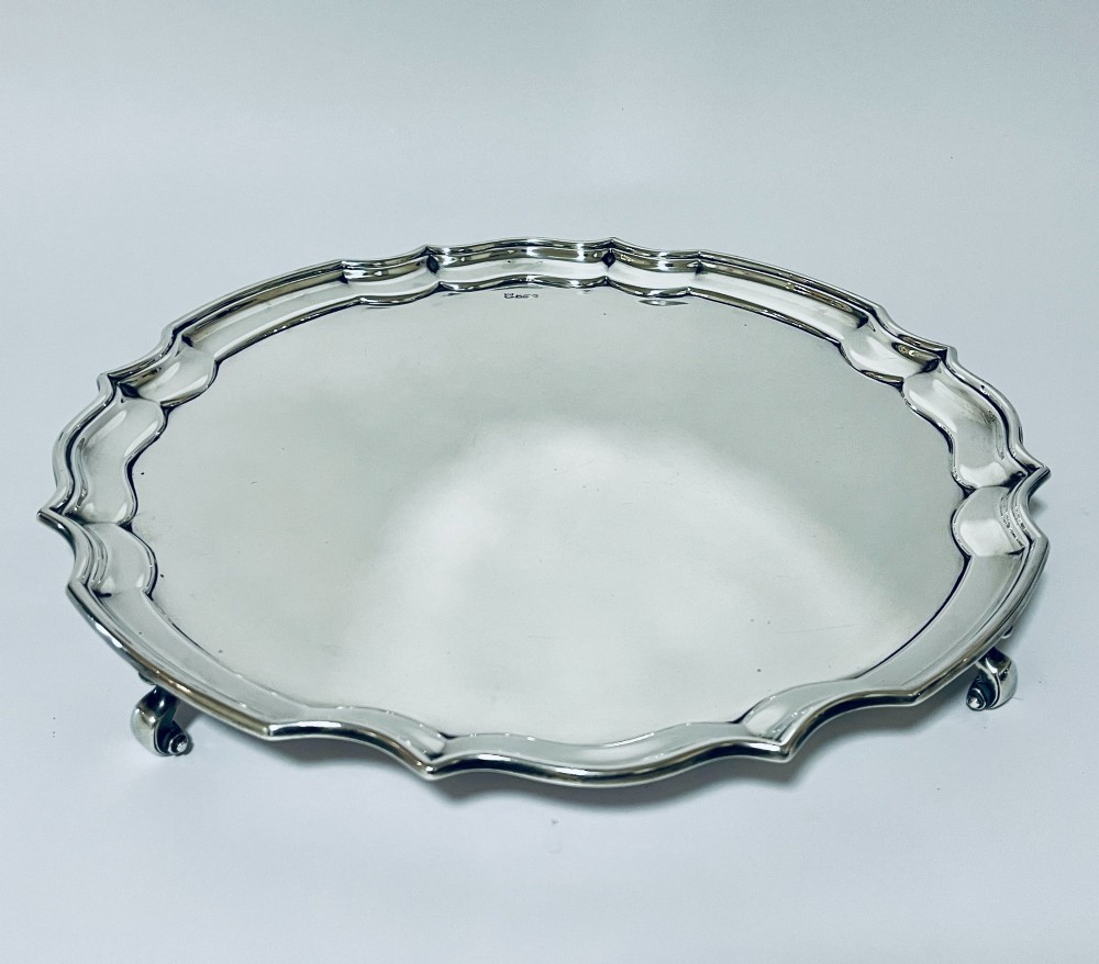 An antique circular silver salver with a moulded piecrust border and raised scroll feet, photographed on a plain white background.