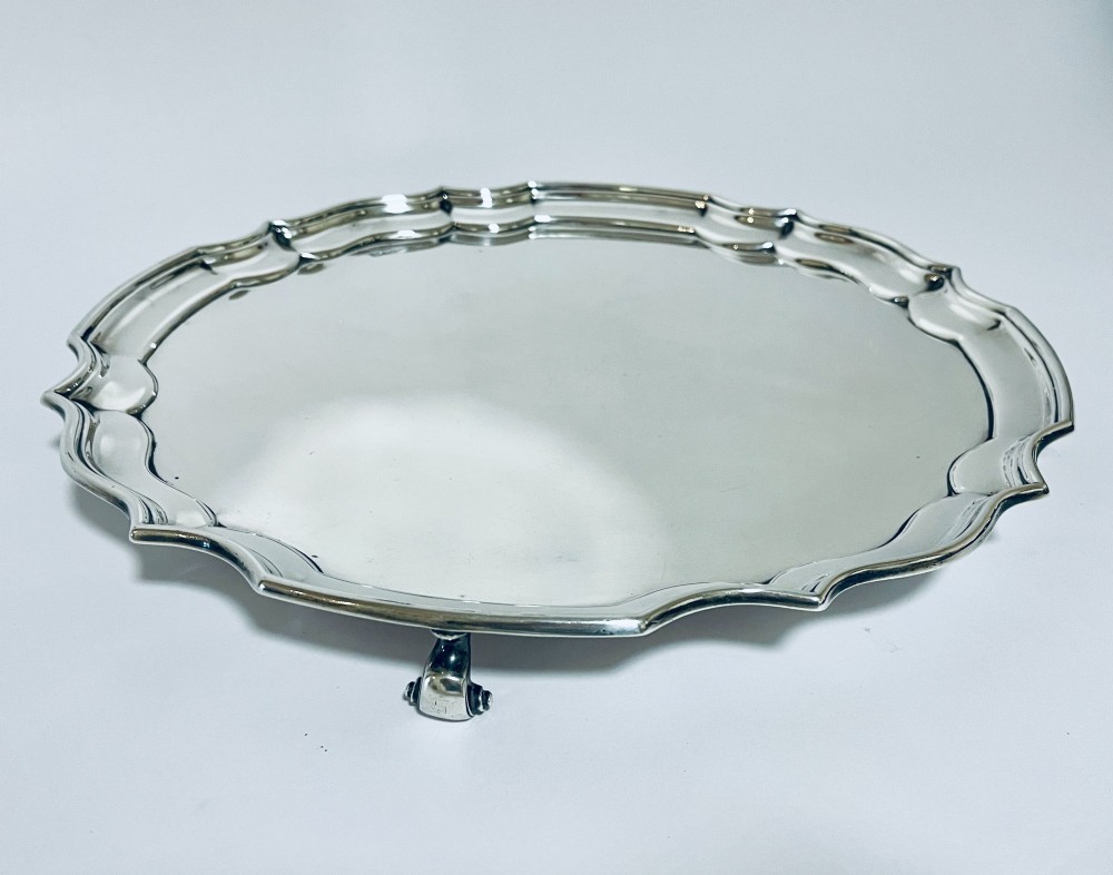 An antique circular silver salver with a moulded piecrust border and raised scroll feet, photographed on a plain white background. View IV