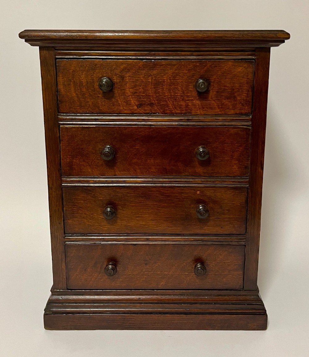 A dark oak miniature tabletop chest of drawers featuring four drawers with turned wooden knobs and a moulded plinth base.