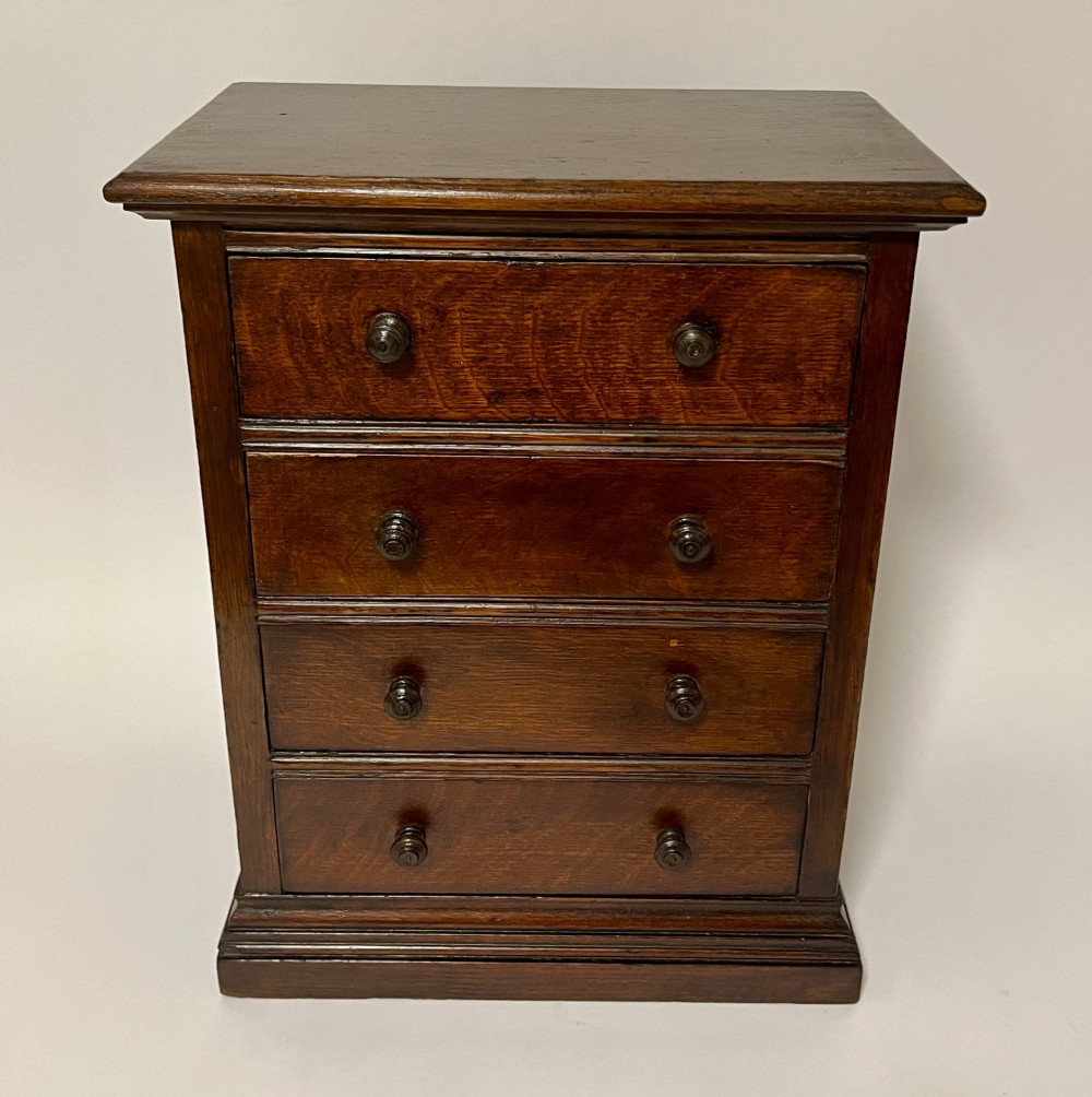 A dark oak miniature tabletop chest of drawers featuring four drawers with turned wooden knobs and a moulded plinth base. View II