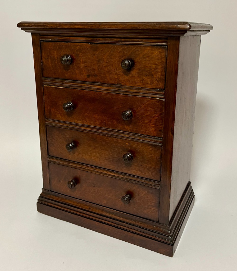 A dark oak miniature tabletop chest of drawers featuring four drawers with turned wooden knobs and a moulded plinth base. View III