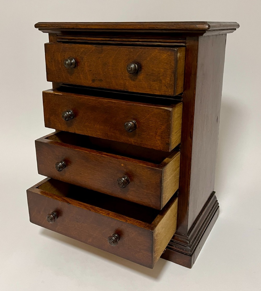 A dark oak miniature tabletop chest of drawers featuring four drawers with turned wooden knobs and a moulded plinth base. View IV