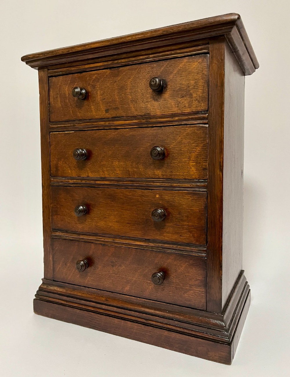 A dark oak miniature tabletop chest of drawers featuring four drawers with turned wooden knobs and a moulded plinth base. View V