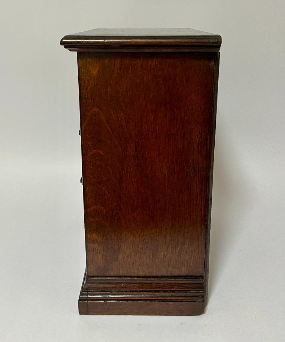 A dark oak miniature tabletop chest of drawers featuring four drawers with turned wooden knobs and a moulded plinth base. View VI
