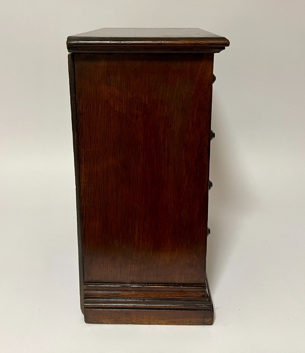 A dark oak miniature tabletop chest of drawers featuring four drawers with turned wooden knobs and a moulded plinth base. View VIII