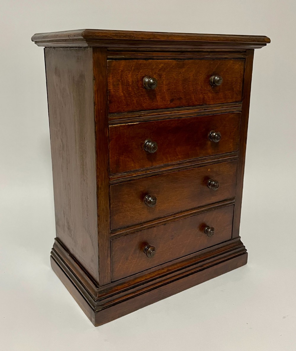 A dark oak miniature tabletop chest of drawers featuring four drawers with turned wooden knobs and a moulded plinth base. View IX