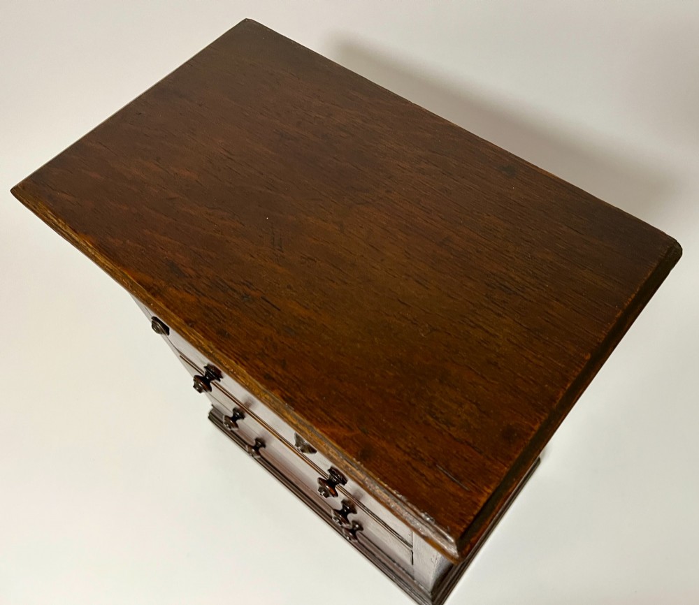 A dark oak miniature tabletop chest of drawers featuring four drawers with turned wooden knobs and a moulded plinth base. View X