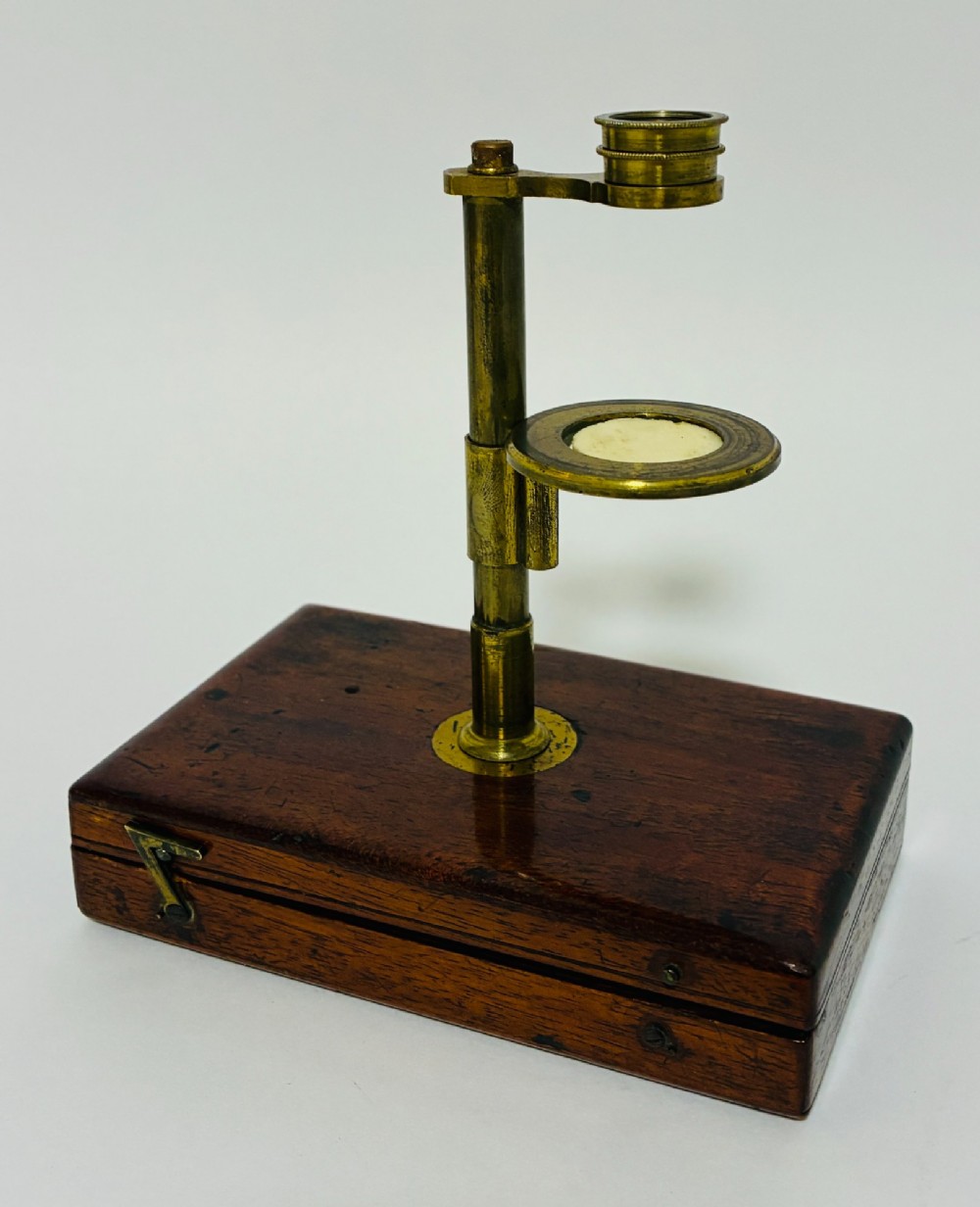 A 19th-century brass botanical field microscope mounted upright on a polished mahogany wooden storage box base.