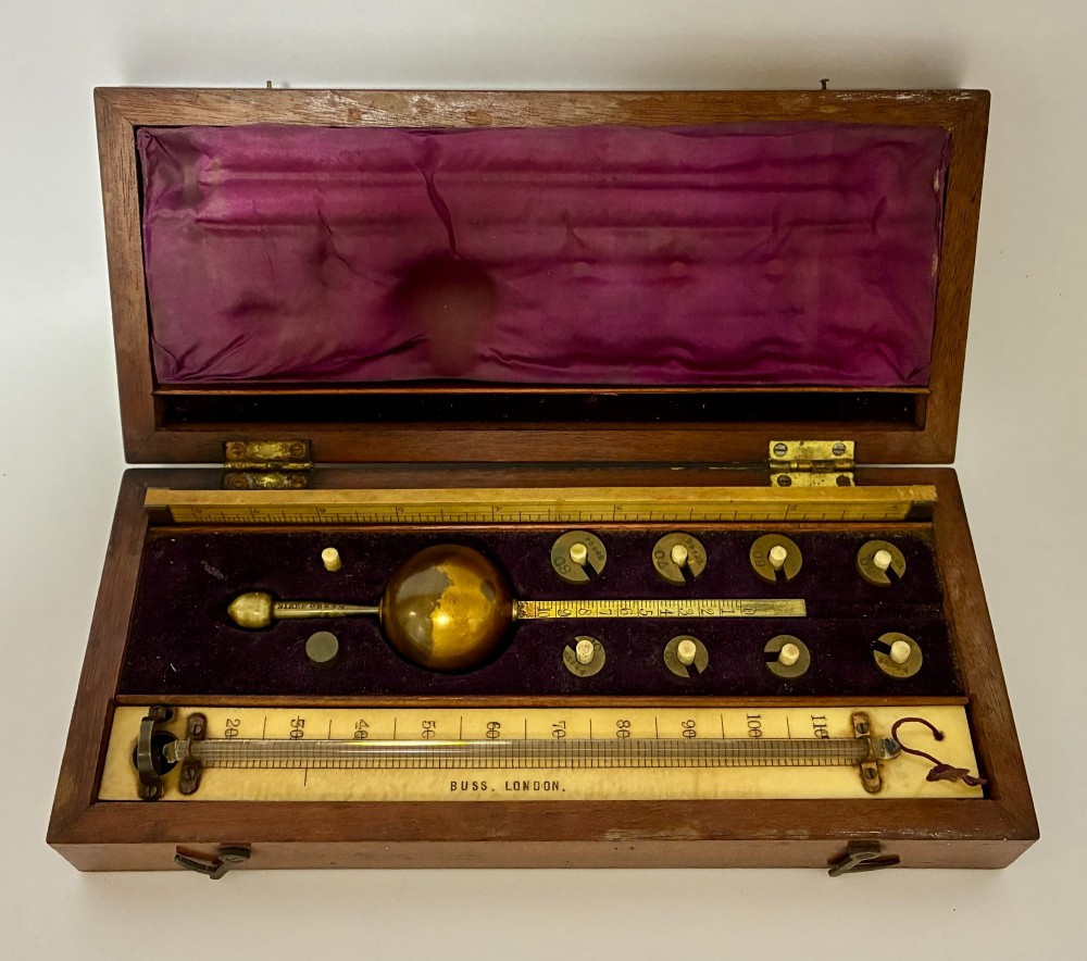 A Victorian Sikes hydrometer set resting inside an open fitted mahogany box lined with purple velvet, containing a brass float, slotted weights, and a thermometer.