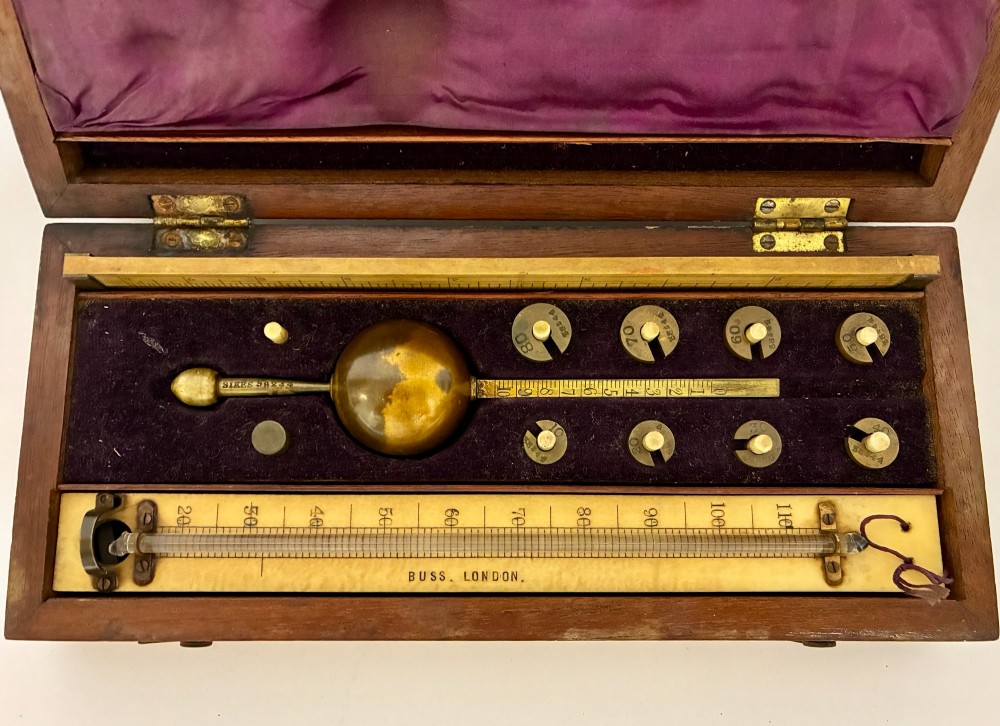 A Victorian Sikes hydrometer set resting inside an open fitted mahogany box lined with purple velvet, containing a brass float, slotted weights, and a thermometer. View II