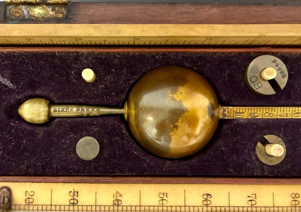 A Victorian Sikes hydrometer set resting inside an open fitted mahogany box lined with purple velvet, containing a brass float, slotted weights, and a thermometer. View III