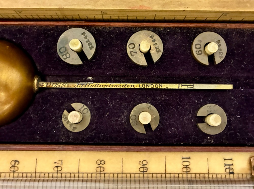 A Victorian Sikes hydrometer set resting inside an open fitted mahogany box lined with purple velvet, containing a brass float, slotted weights, and a thermometer. View IV