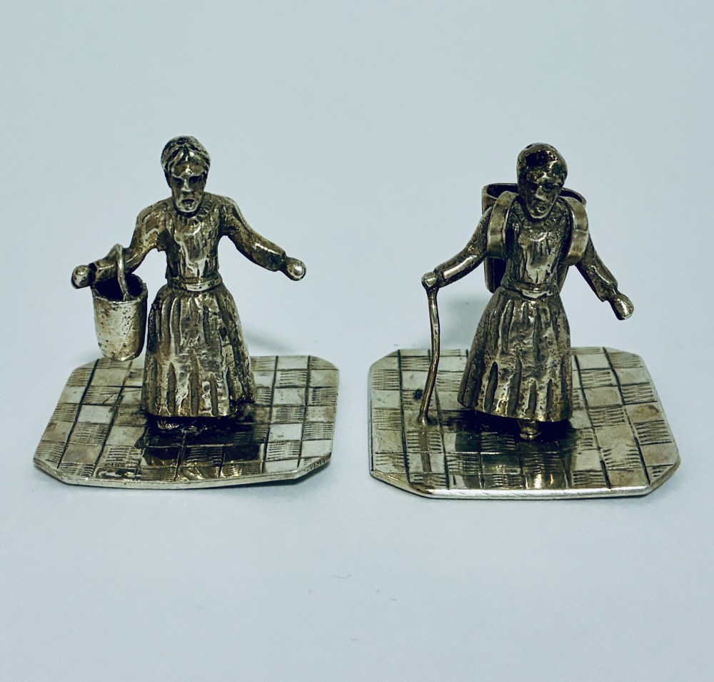 A pair of miniature silver figures depicting figures with a bucket and a walking stick on square patterned bases.