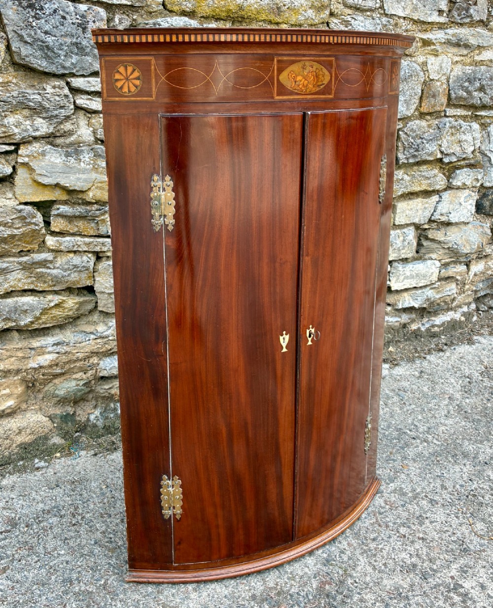 A Georgian bow-front mahogany corner cupboard featuring detailed marquetry inlays, decorative brass hinges, and central keyhole escutcheons. View II