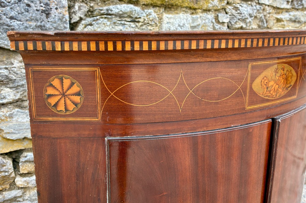 A Georgian bow-front mahogany corner cupboard featuring detailed marquetry inlays, decorative brass hinges, and central keyhole escutcheons. View III