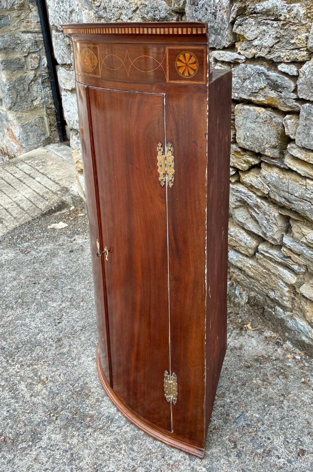 A Georgian bow-front mahogany corner cupboard featuring detailed marquetry inlays, decorative brass hinges, and central keyhole escutcheons. View V