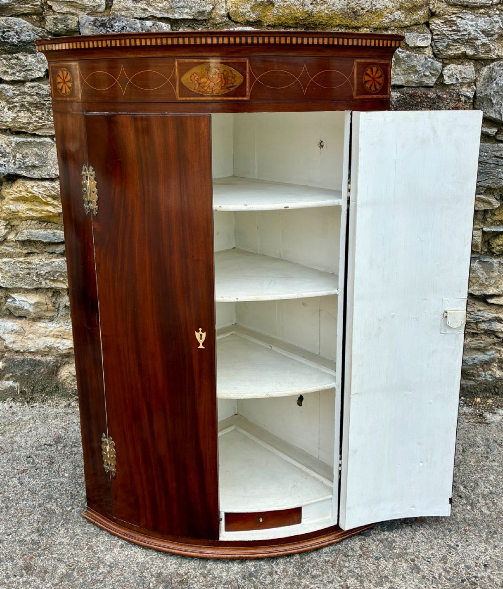 A Georgian bow-front mahogany corner cupboard featuring detailed marquetry inlays, decorative brass hinges, and central keyhole escutcheons. View X