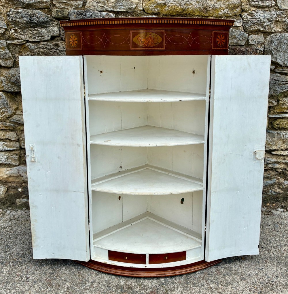 A Georgian bow-front mahogany corner cupboard featuring detailed marquetry inlays, decorative brass hinges, and central keyhole escutcheons. View XII