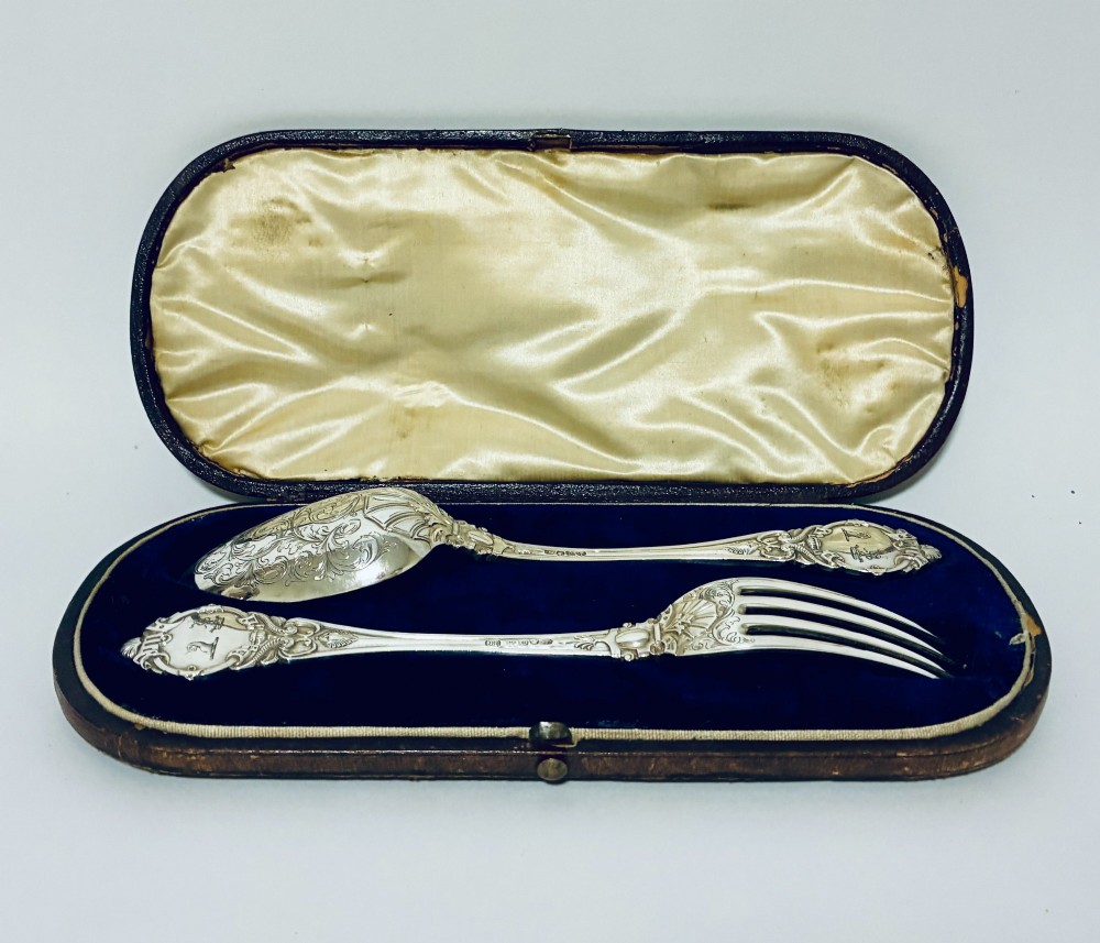 An antique Victorian silver christening spoon and fork with intricate foliate engraving, arranged inside a velvet-lined dark leather presentation case.
