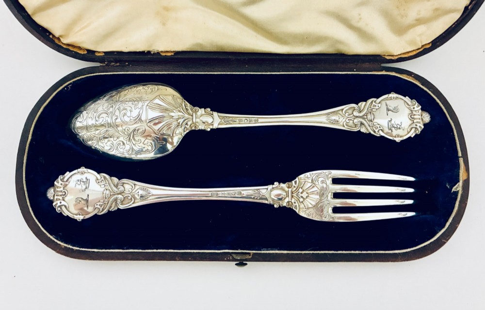 An antique Victorian silver christening spoon and fork with intricate foliate engraving, arranged inside a velvet-lined dark leather presentation case. View II
