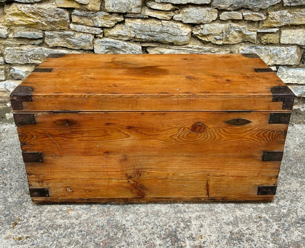 An antique rectangular pine campaign trunk with dark metal corner brackets, a brass escutcheon, and visible natural wood grain.