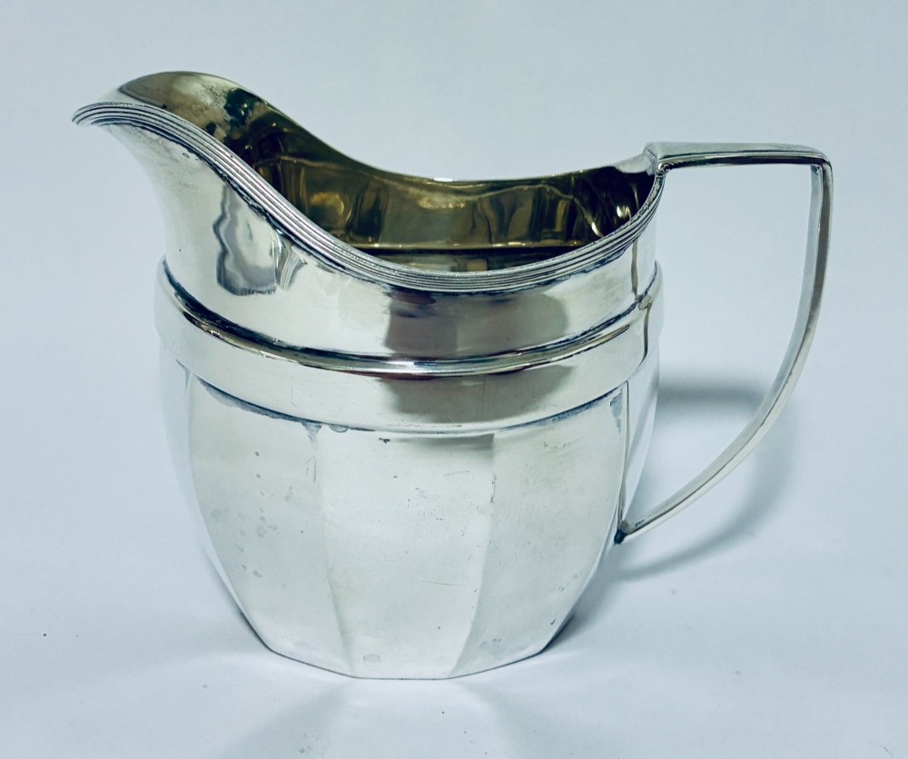 An antique polished silver milk jug featuring a faceted lower body, a shaped pouring spout with reeded rim, and an angular handle.