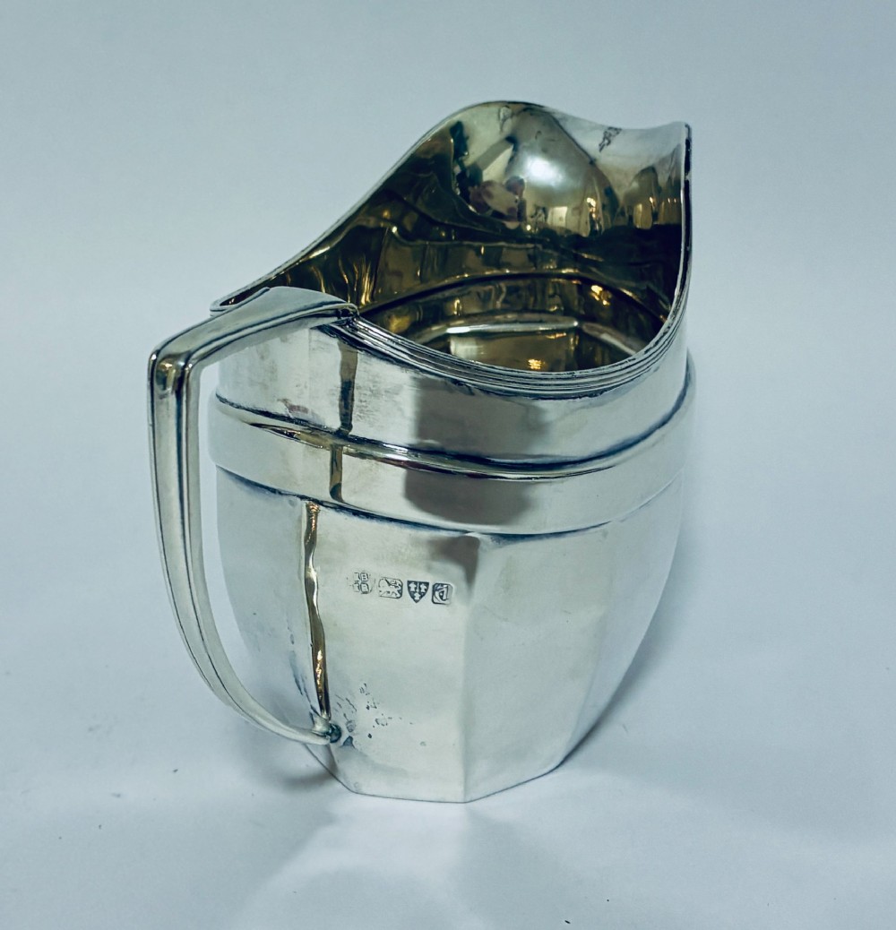 An antique polished silver milk jug featuring a faceted lower body, a shaped pouring spout with reeded rim, and an angular handle. View V