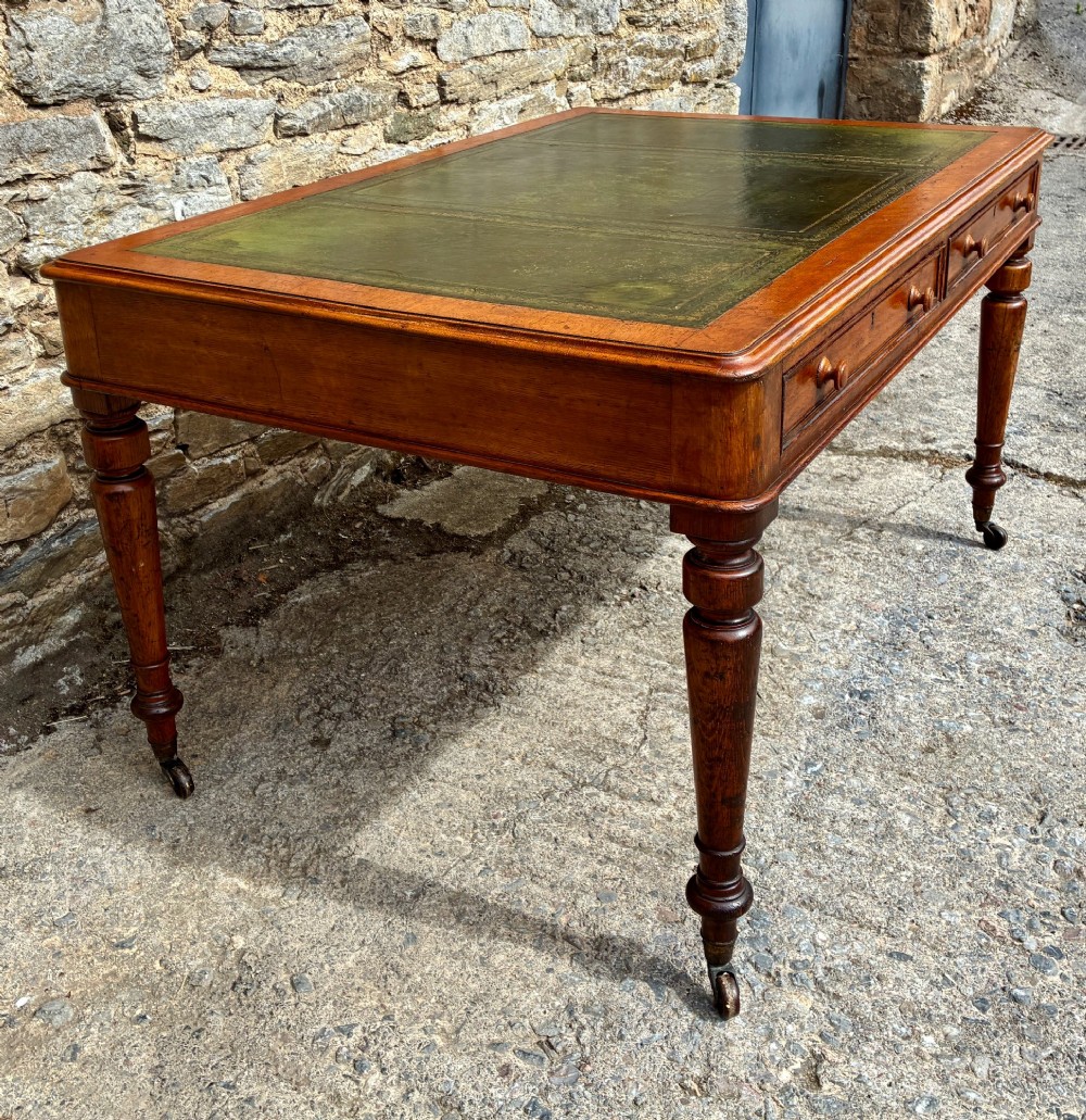 A Victorian oak writing desk featuring a green leather insert top, frieze drawers with wooden turned knobs, and turned legs with brass castors. View IV