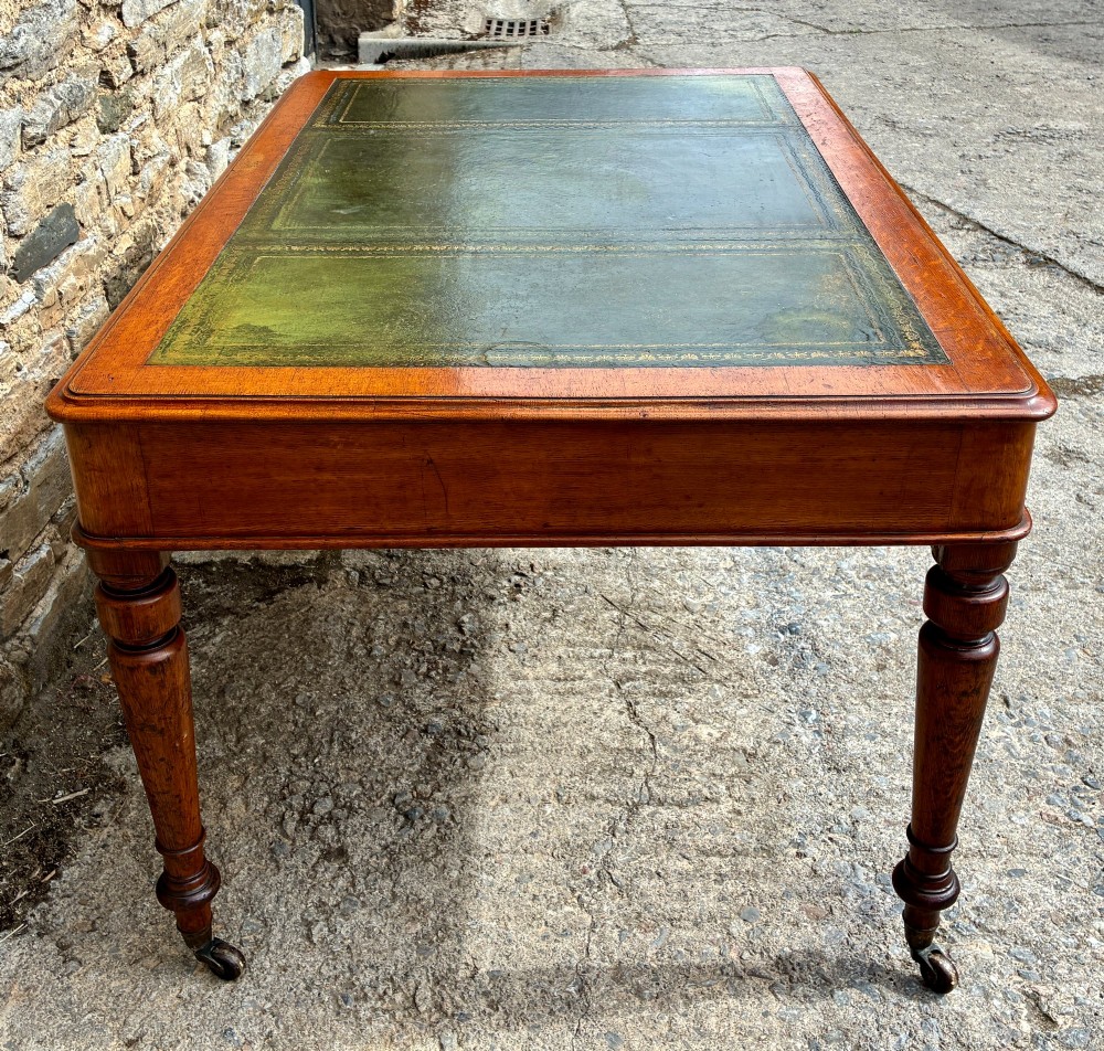 A Victorian oak writing desk featuring a green leather insert top, frieze drawers with wooden turned knobs, and turned legs with brass castors. View VI