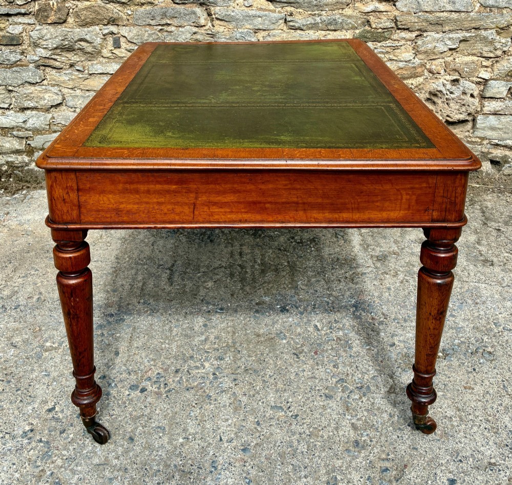 A Victorian oak writing desk featuring a green leather insert top, frieze drawers with wooden turned knobs, and turned legs with brass castors. View XIV