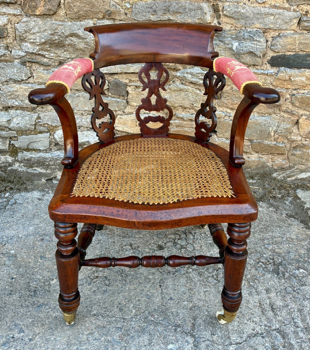 A Victorian mahogany desk chair featuring a curved top rail, pierced decorative splats, fabric-wrapped armrests, a woven cane seat, and turned legs with brass castors.